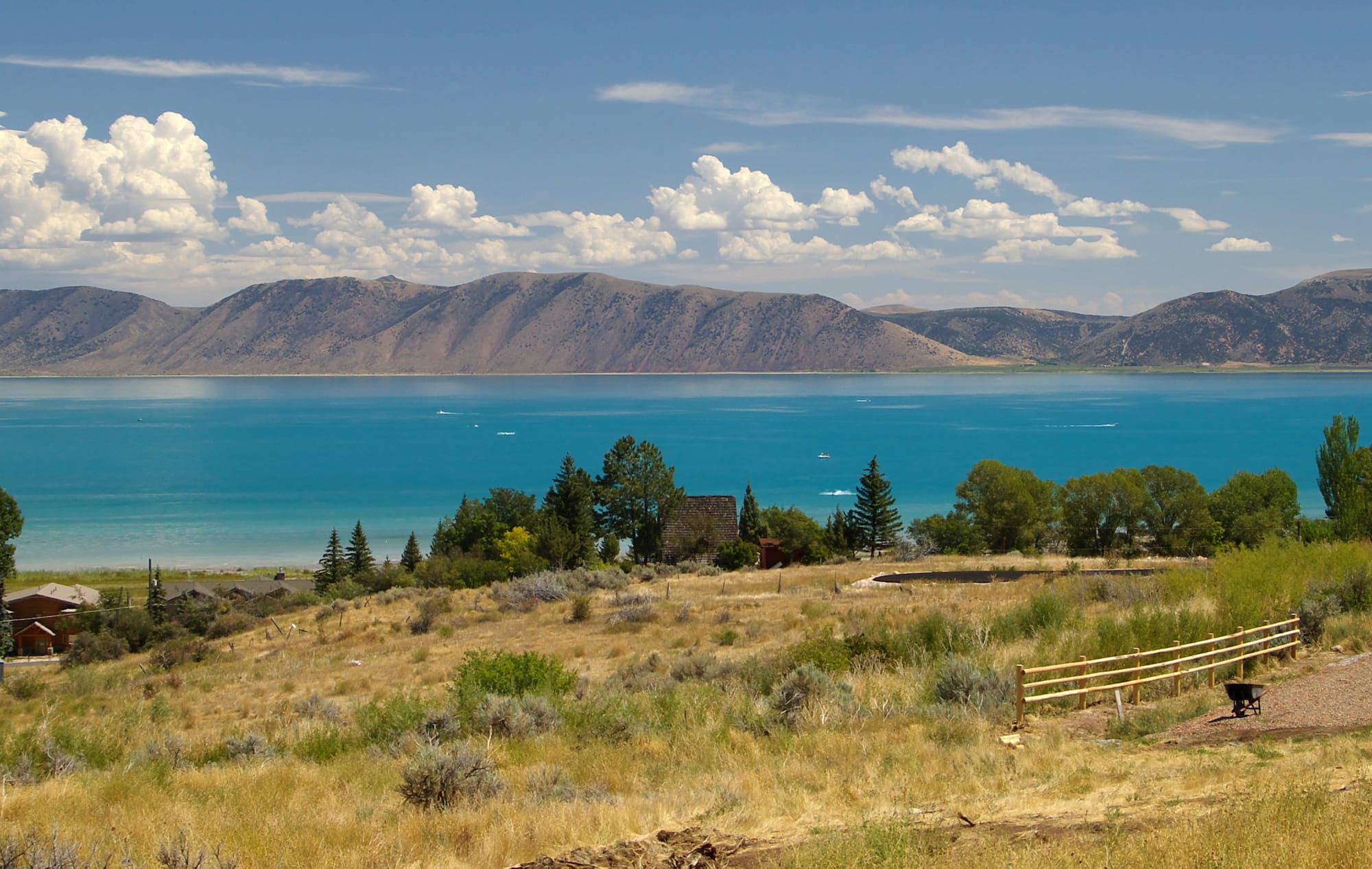 Turquoise Bear Lake below dry, sage-covered hills, with a split-rail fence and trees in the foreground