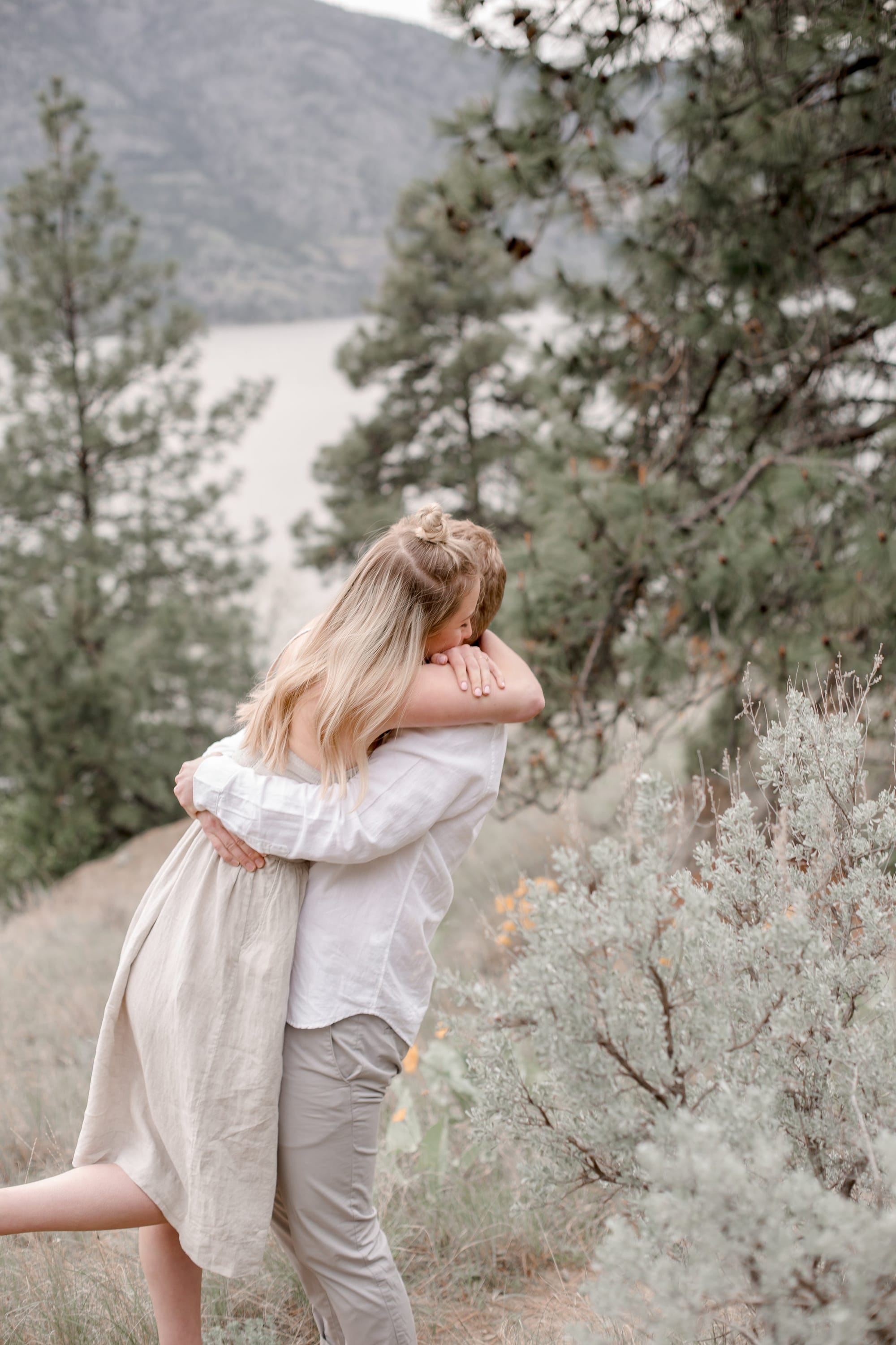 Two people hugging on a hillside among sagebrush and pines, with a lake and mountains behind them